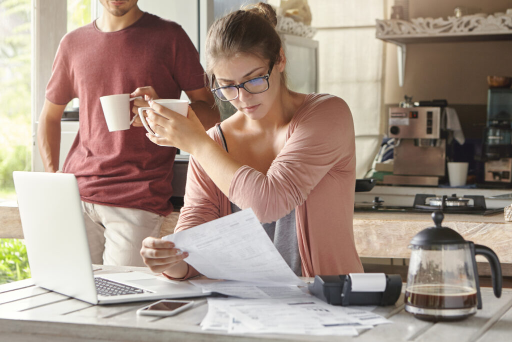 young caucasian family calculating bills, reviewing finances and planning family budget together in kitchen: wife studying document, asking her husband in background to pour some more coffee